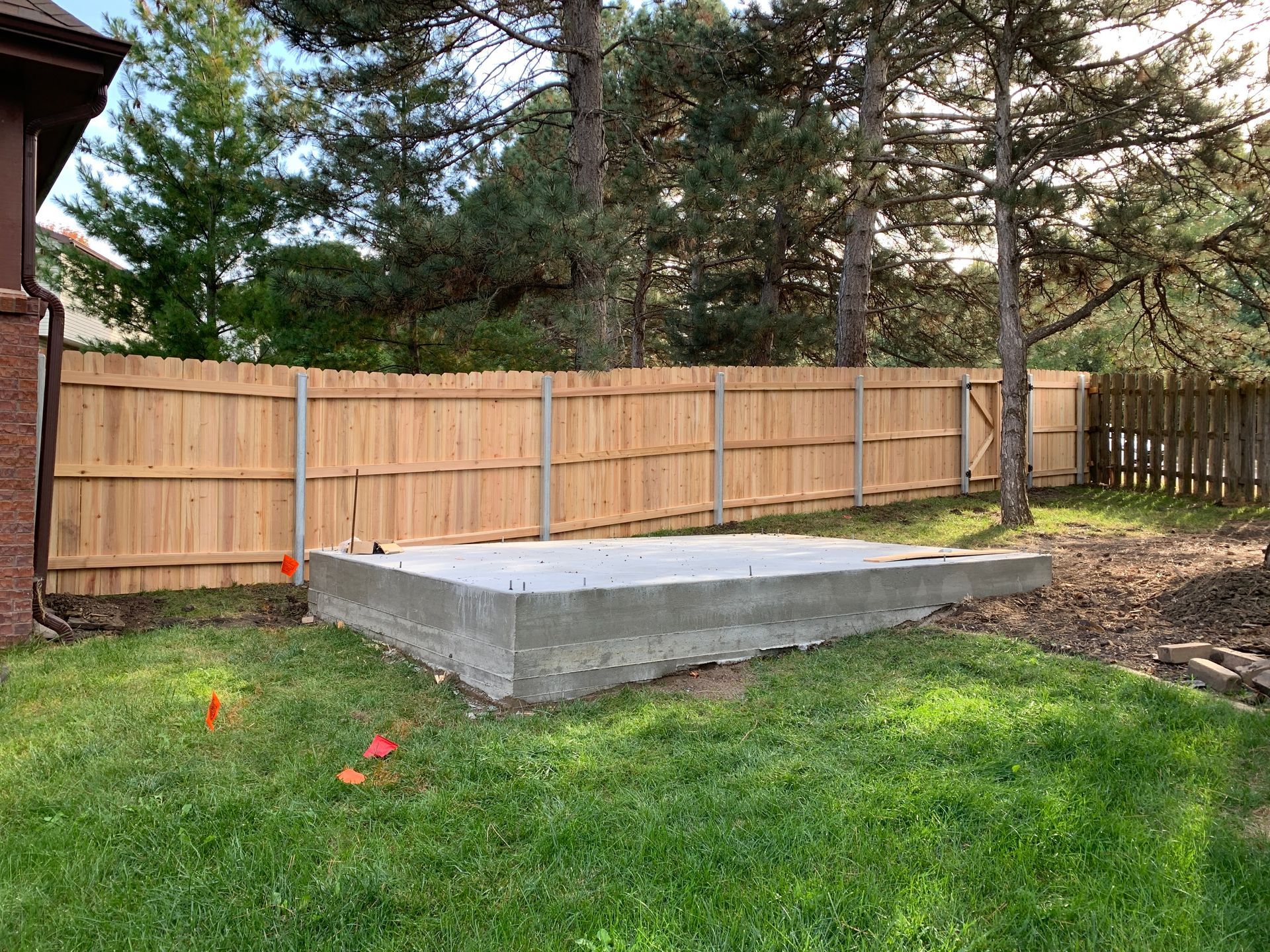 A concrete pad sits in a grassy backyard, near a wooden fence. Trees and a house are in the background.