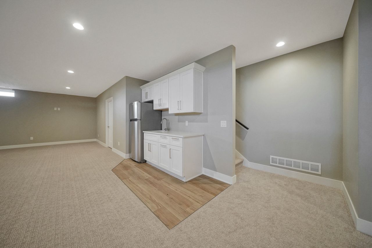 A finished basement with beige carpet, a vinyl plank patch under a white shaker kitchenette with a stainless refrigerator, and a stair run at the right.