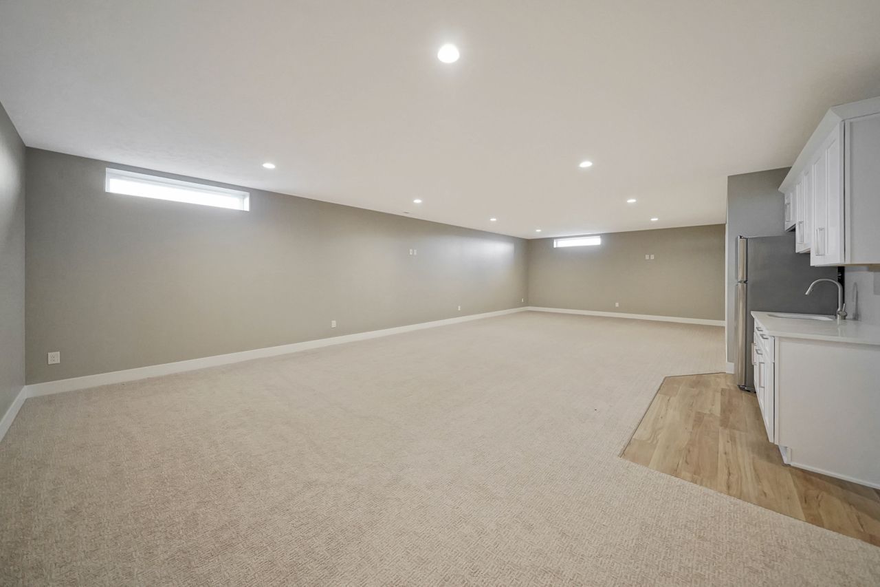 A large finished basement room with gray walls, beige carpet, high narrow windows, recessed lights and a white kitchenette at the right.