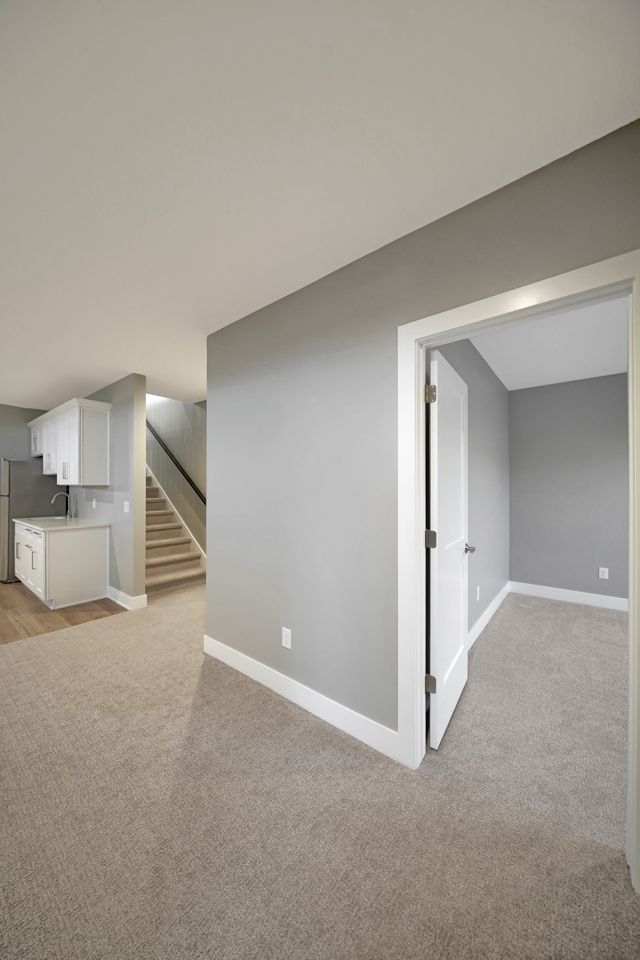 A basement with beige carpet, gray walls, a white kitchenette with a stainless refrigerator, a staircase and an open door to a carpeted room.