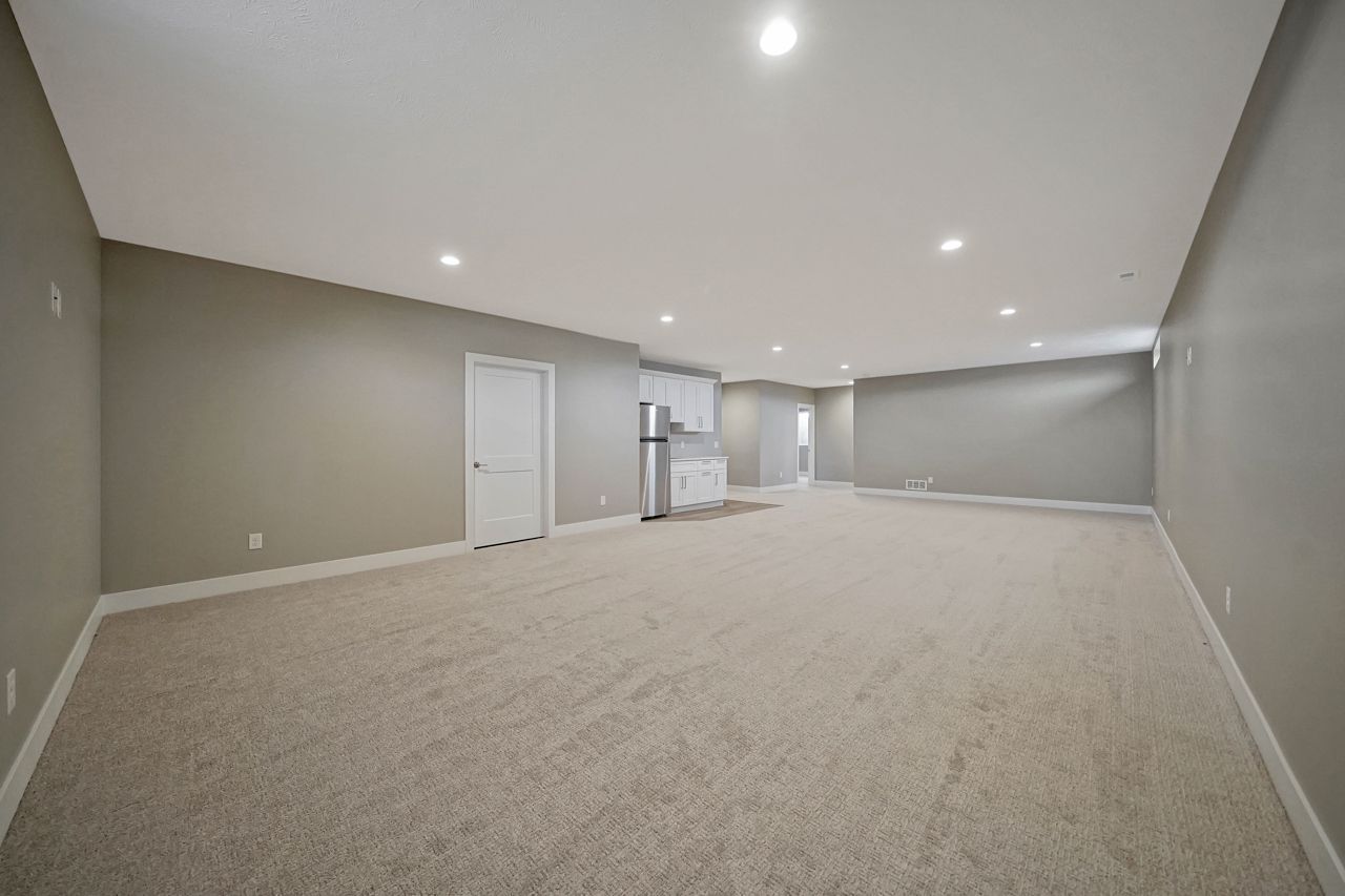 A large carpeted room with gray walls, recessed lighting, a white door and a small kitchenette with white cabinets and a stainless refrigerator.