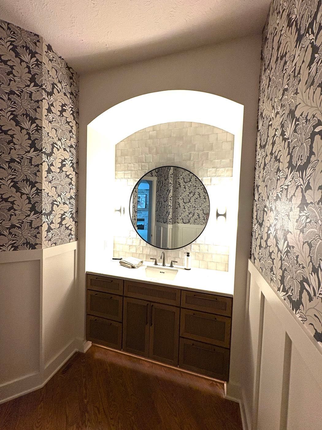 A bathroom vanity nook with a round mirror, tiled backsplash and floral wallpaper accent walls, Lincoln, NE.