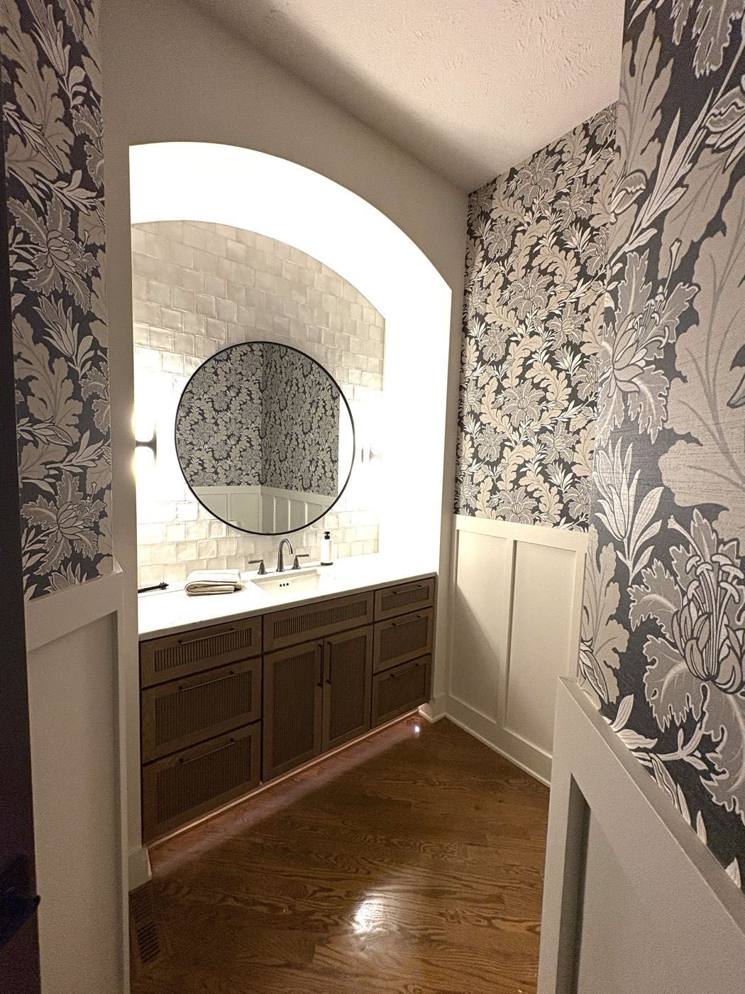 A bathroom hallway leading to a tiled vanity nook with a round mirror and floral wallpaper, Lincoln, NE.