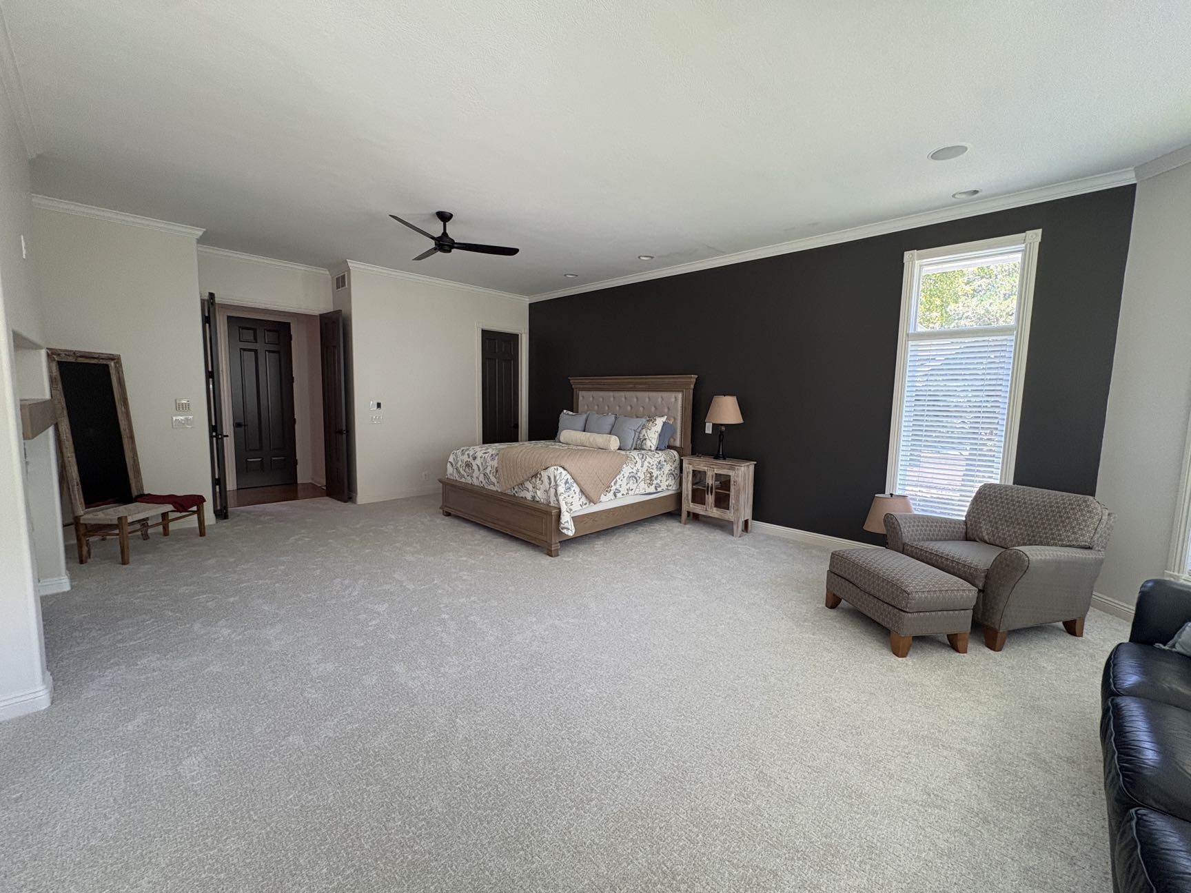 A large bedroom with a dark accent wall, a tufted-headboard bed, a glass-front nightstand, an armchair with ottoman and a leaning mirror, Lincoln, NE.