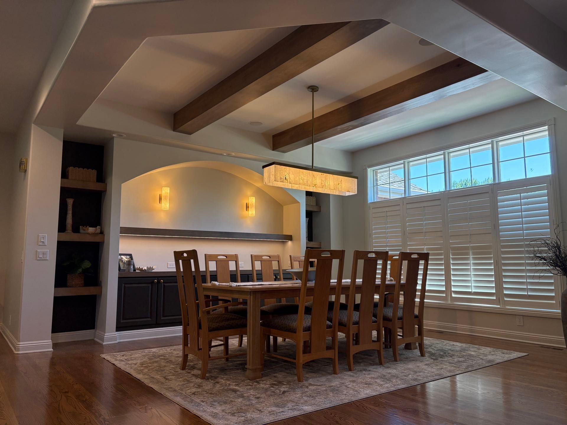 A dining room with a wood beam ceiling, a long wood table seating eight, and an arched built-in bar niche, Lincoln, NE.