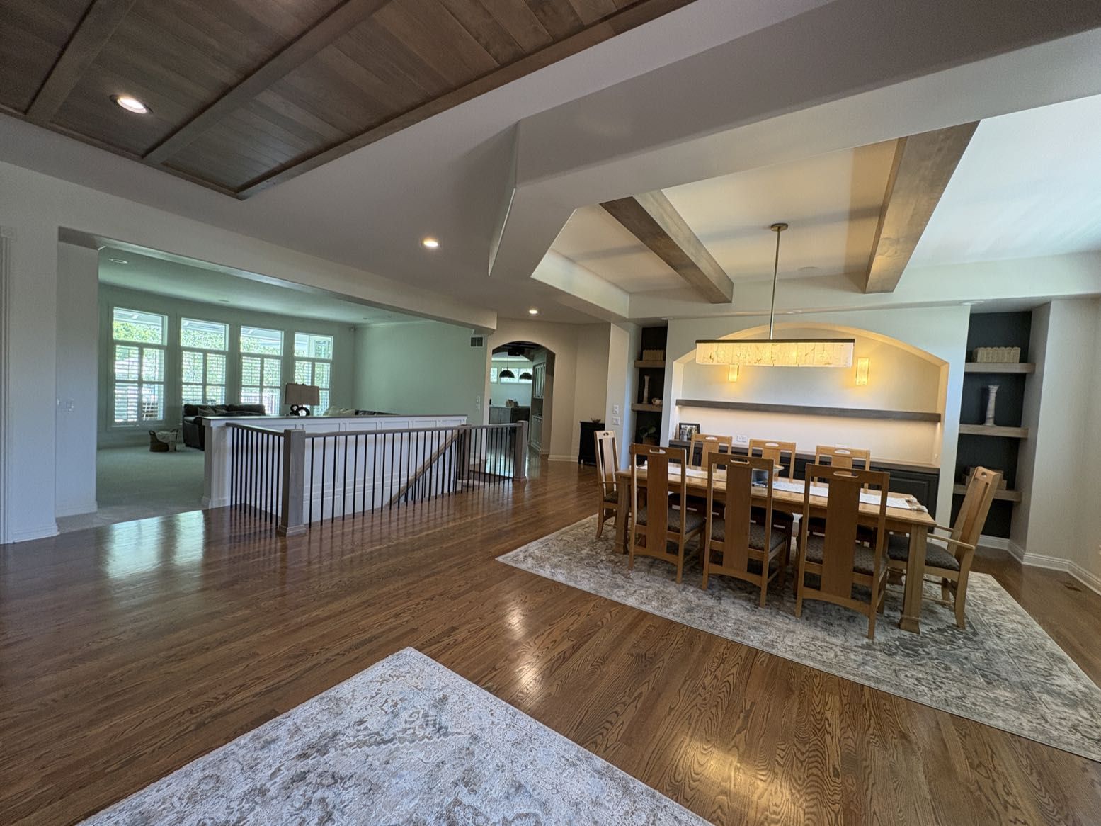A dining room with a wood beam ceiling and long table, seen across a railed stairwell opening, with the great room at left, Lincoln, NE.