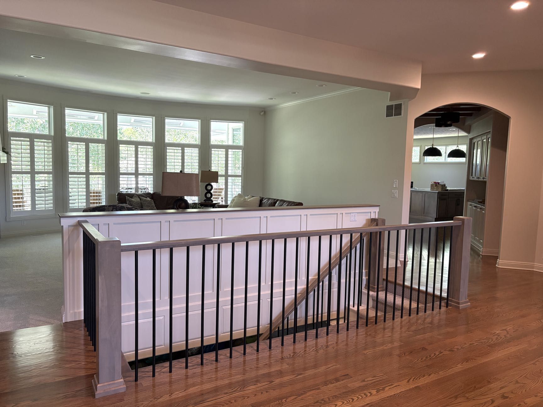 An open great room with shuttered windows and a sofa seen across a railed stairwell opening, with the kitchen through an arch, Lincoln, NE.