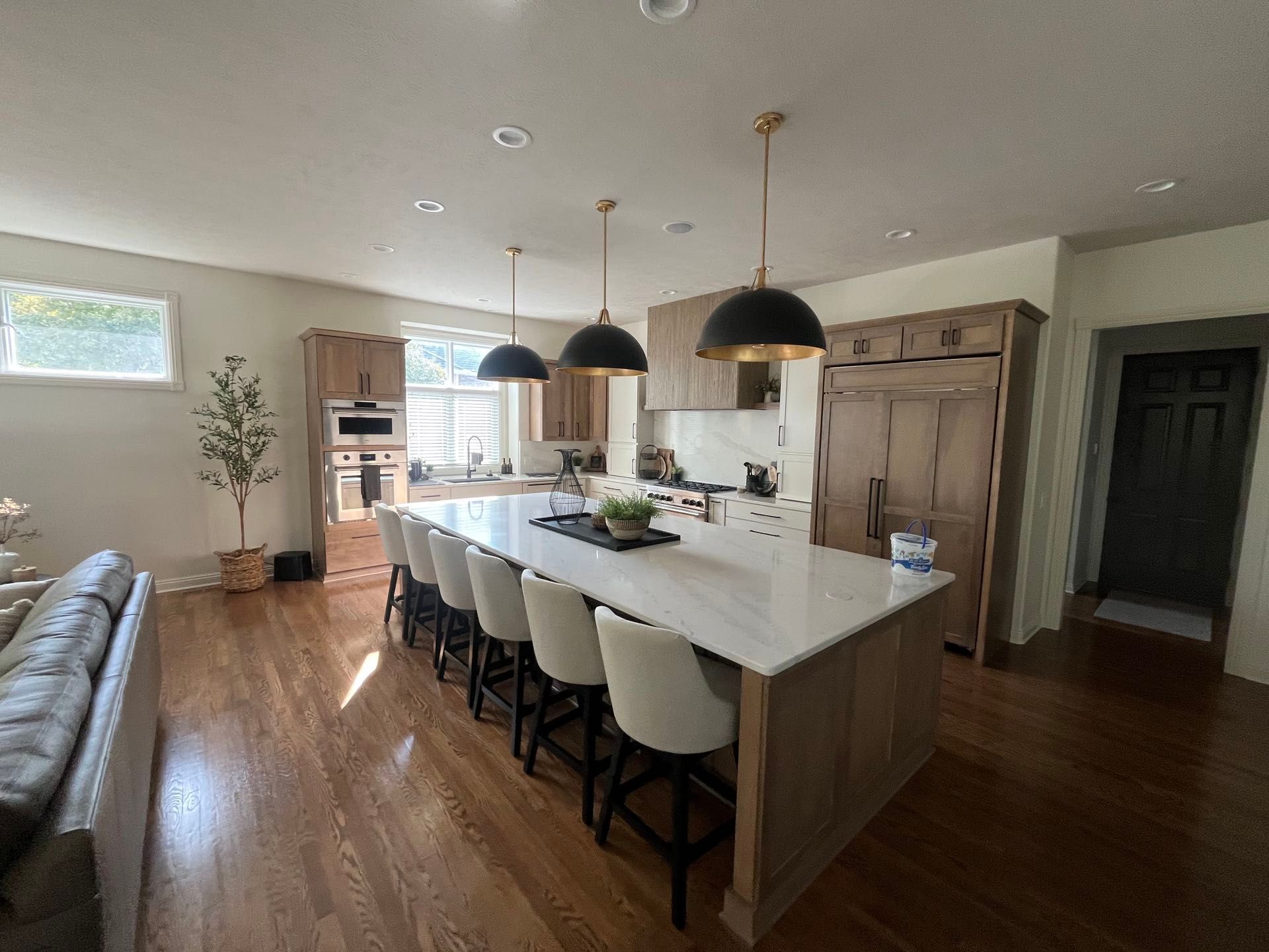 A kitchen with a long white quartz island, light upholstered barstools, black dome pendant lights and stained wood cabinetry, Lincoln, NE.