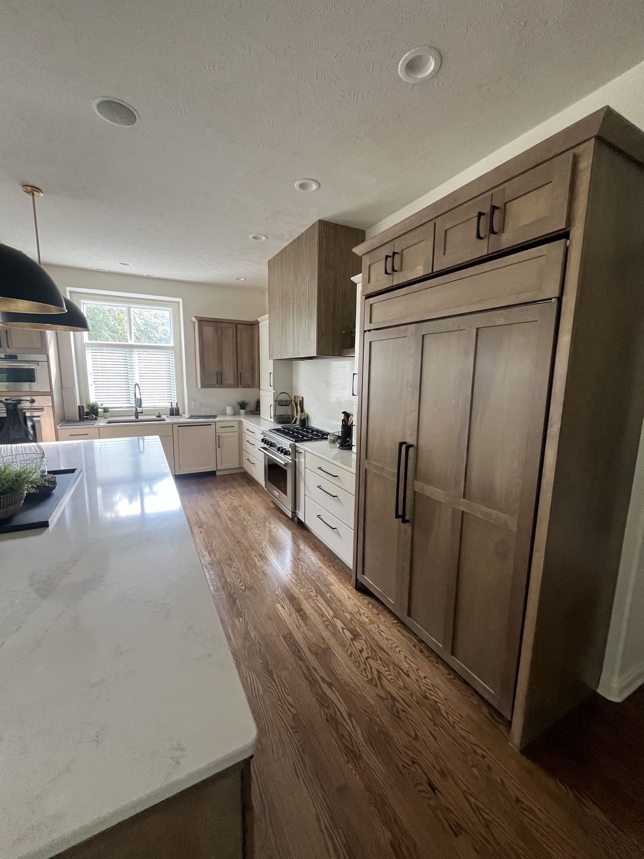 A kitchen seen past a quartz island to white cabinetry, a range under a wood hood, and a stained wood paneled refrigerator wall, Lincoln, NE.