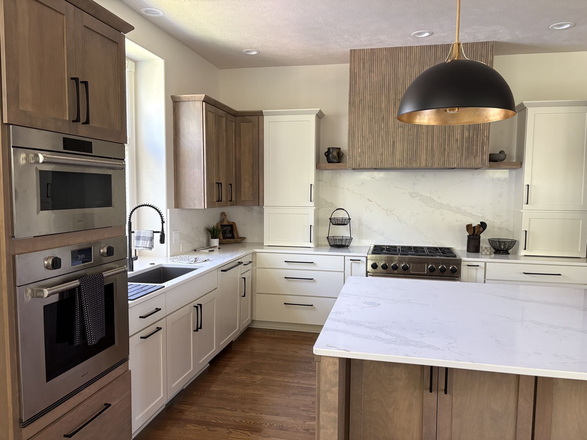 A kitchen wall with white and stained wood cabinetry, a wood-paneled range hood, stainless double ovens and gas cooktop, and quartz counters, Lincoln, NE.