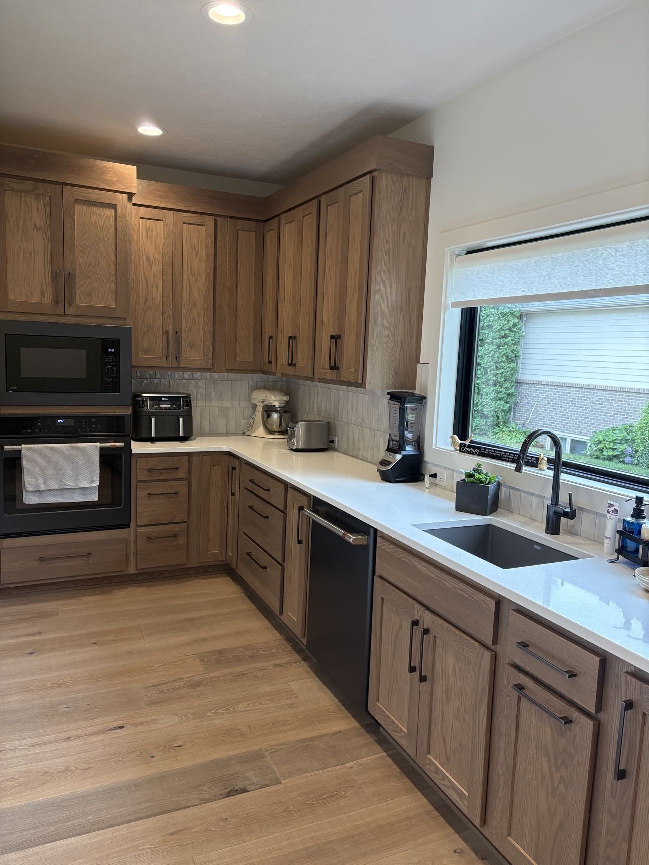 A kitchen with stained oak cabinetry, black built-in microwave and wall oven, black dishwasher, quartz counters and a sink under a window, Lincoln, NE.