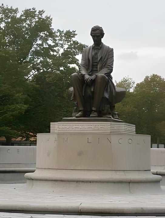 Statue of Abraham Lincoln seated on a pedestal in a park, with trees and overcast sky behind it