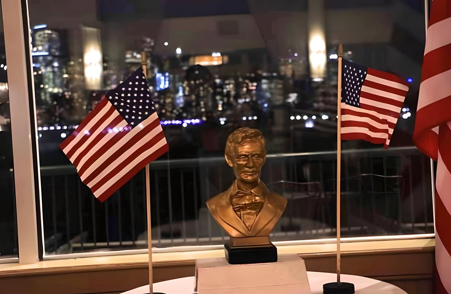 Bronze statue of Abraham Lincoln on a pedestal between two American flags, lit against a nighttime backdrop of jersey city