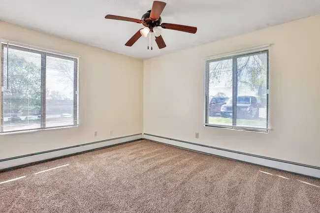 Empty beige bedroom with carpet, two windows with blinds, and a ceiling fan.
