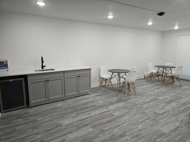 A gray-floored room with white walls, a small kitchenette, and two tables with white chairs.