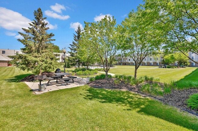 Lush green park with picnic tables, grills, trees, and buildings under a blue sky with clouds.