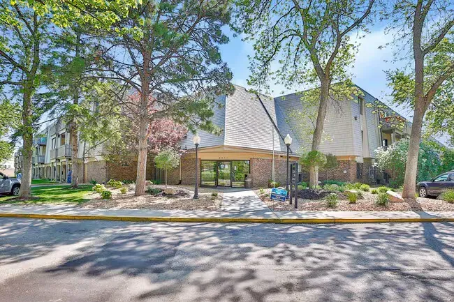 Apartment building exterior with trees, brick entrance, and parked cars on a sunny day.