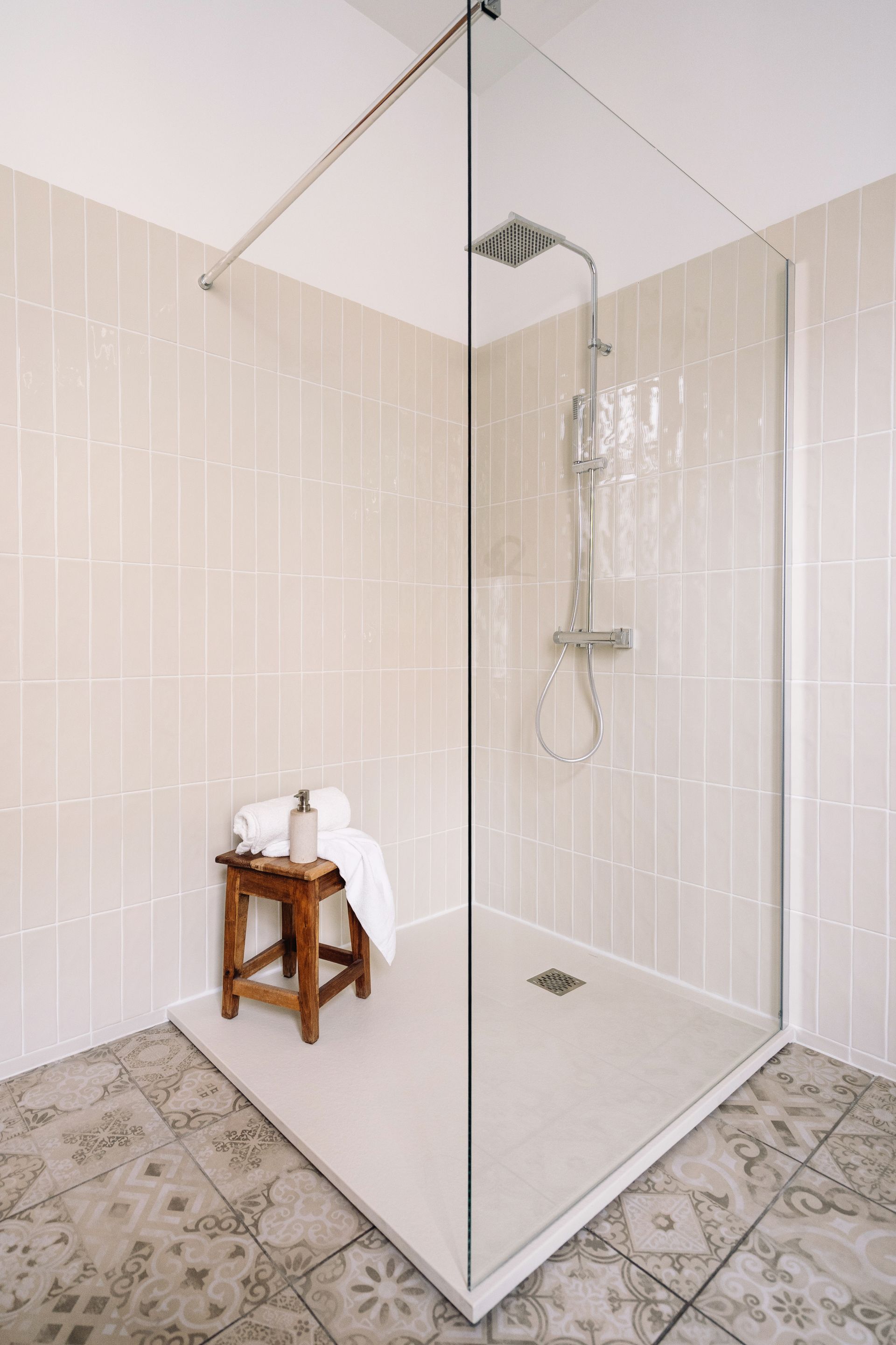 A walk-in shower with light tiles, a glass screen, an overhead shower head, and a wooden stool with a white towel.