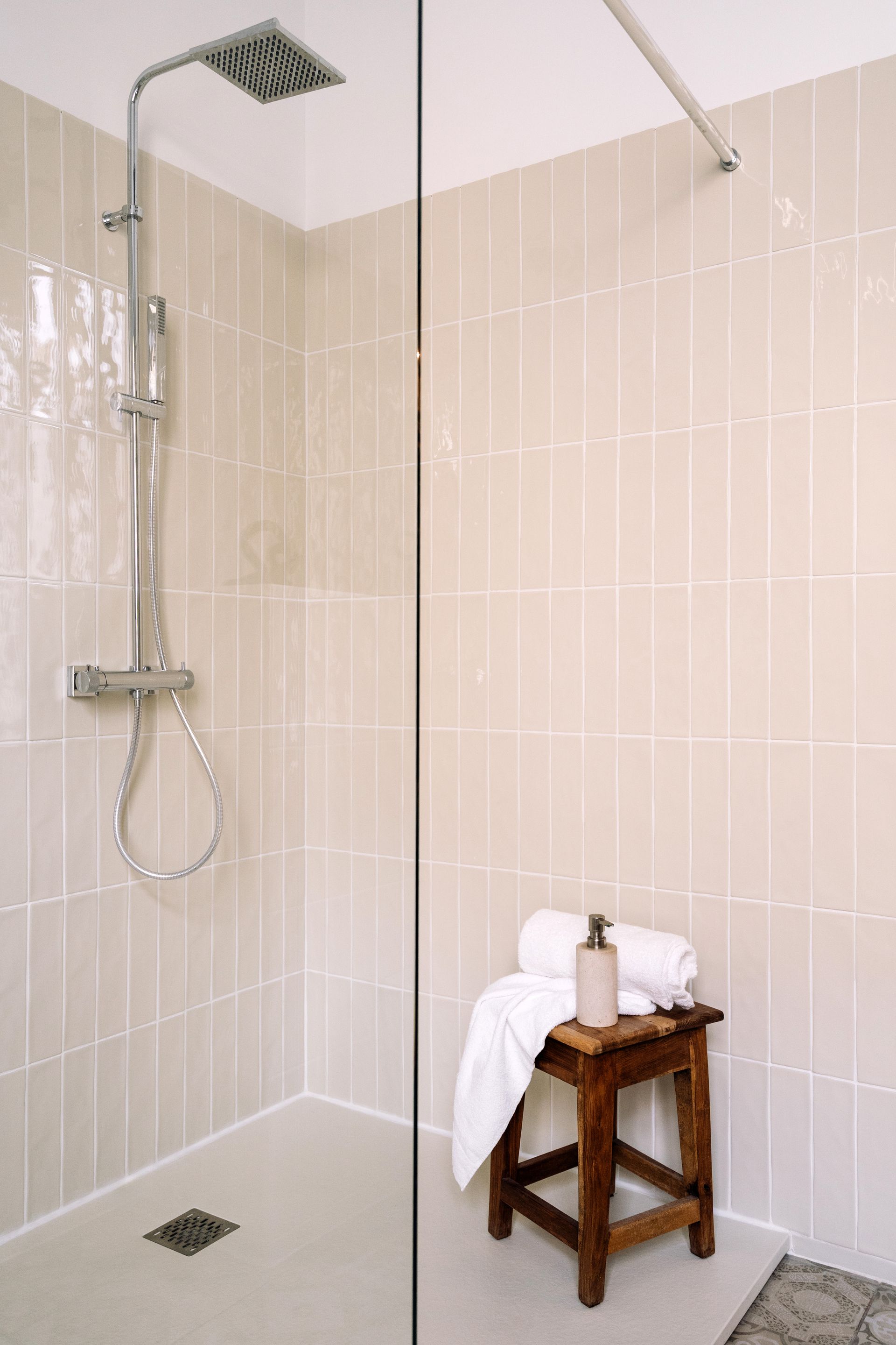 A shower stall with light beige vertical wall tiles, a chrome rain shower head, and a wooden stool with a white towel.