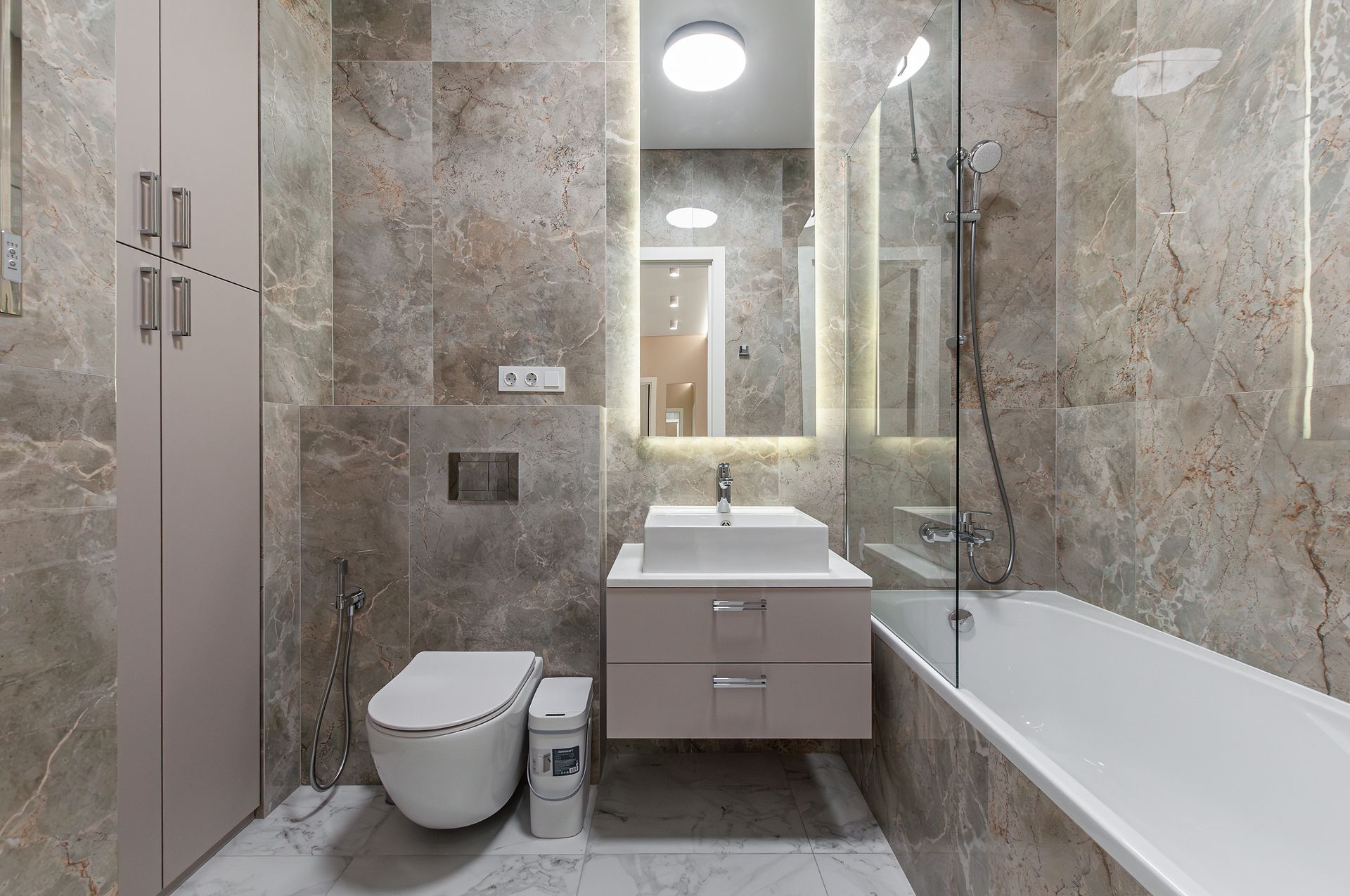 A modern bathroom with marble-tiled walls, a wall-mounted vanity and toilet, and a glass-enclosed bathtub.