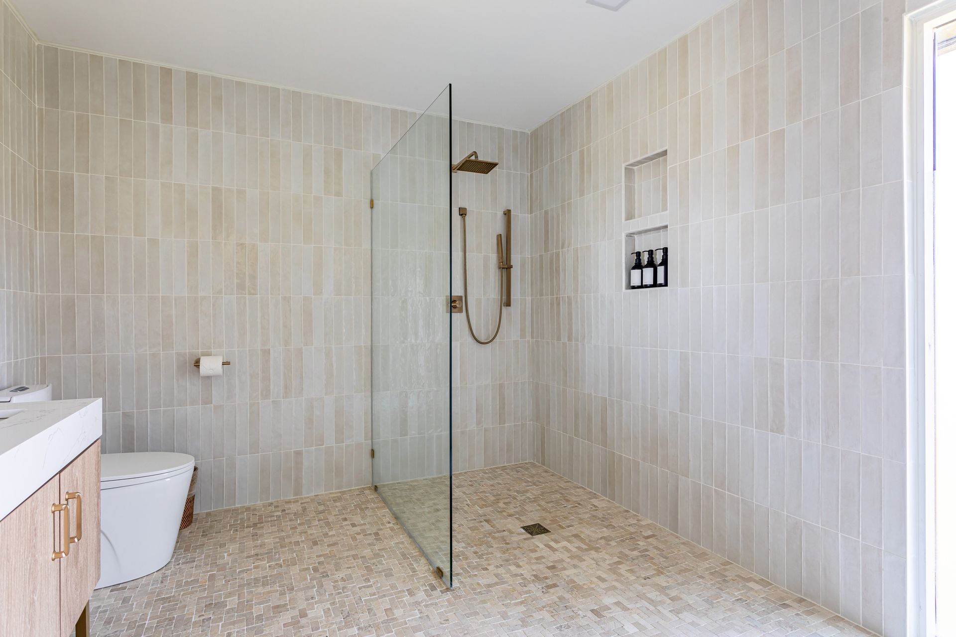A modern bathroom with light, vertical wall tiles, a pebble floor, a glass-enclosed shower, and a wooden vanity.