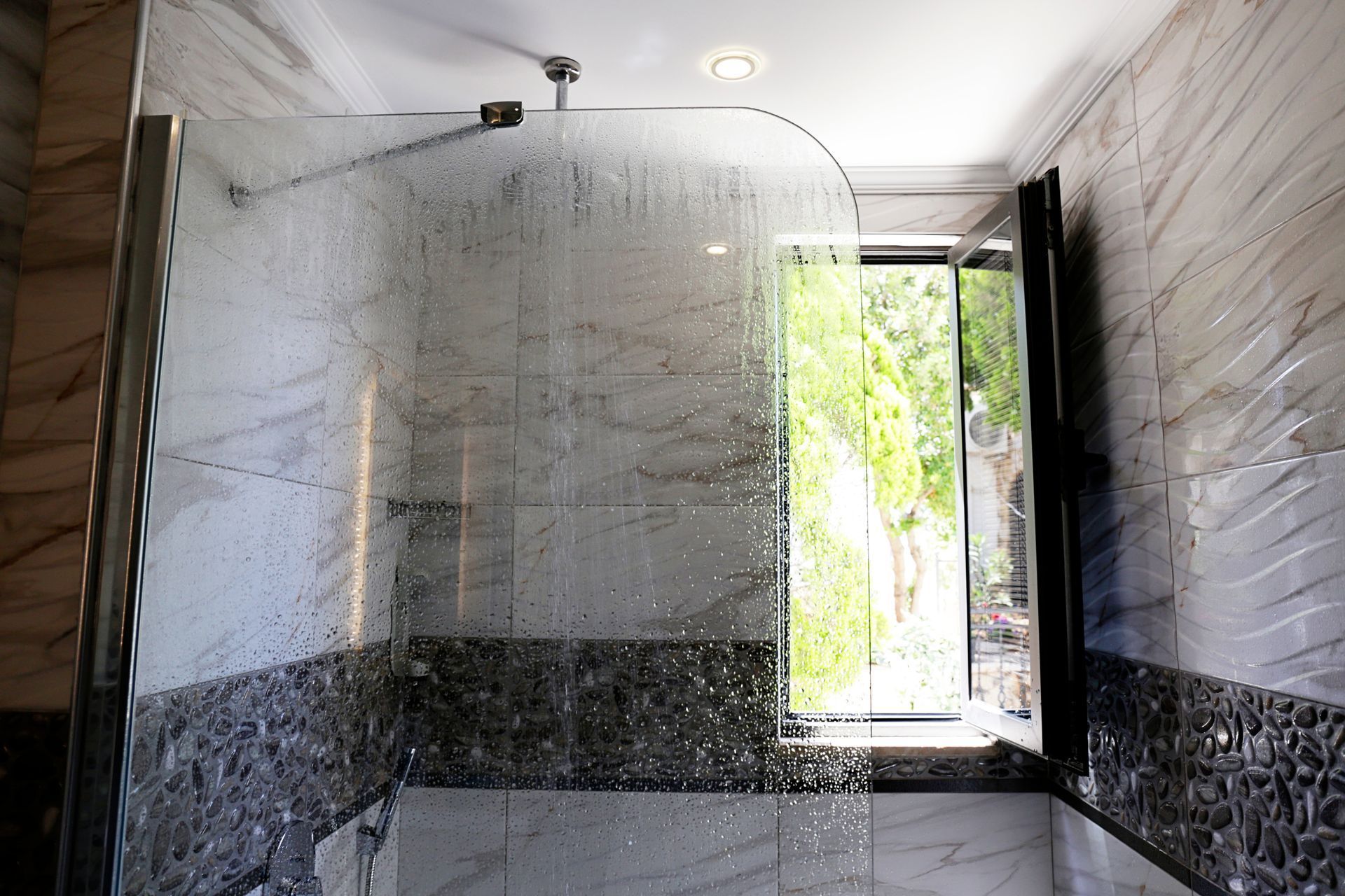 A shower stall with light marbled tile, a decorative stone border, and a glass screen, positioned by a window with trees.
