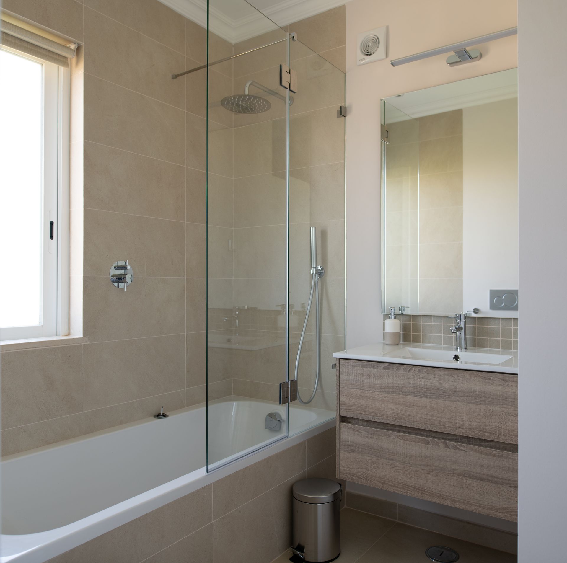 A modern bathroom with a white bathtub, a glass shower screen, light beige wall tiles, and a wood-veneer vanity unit.