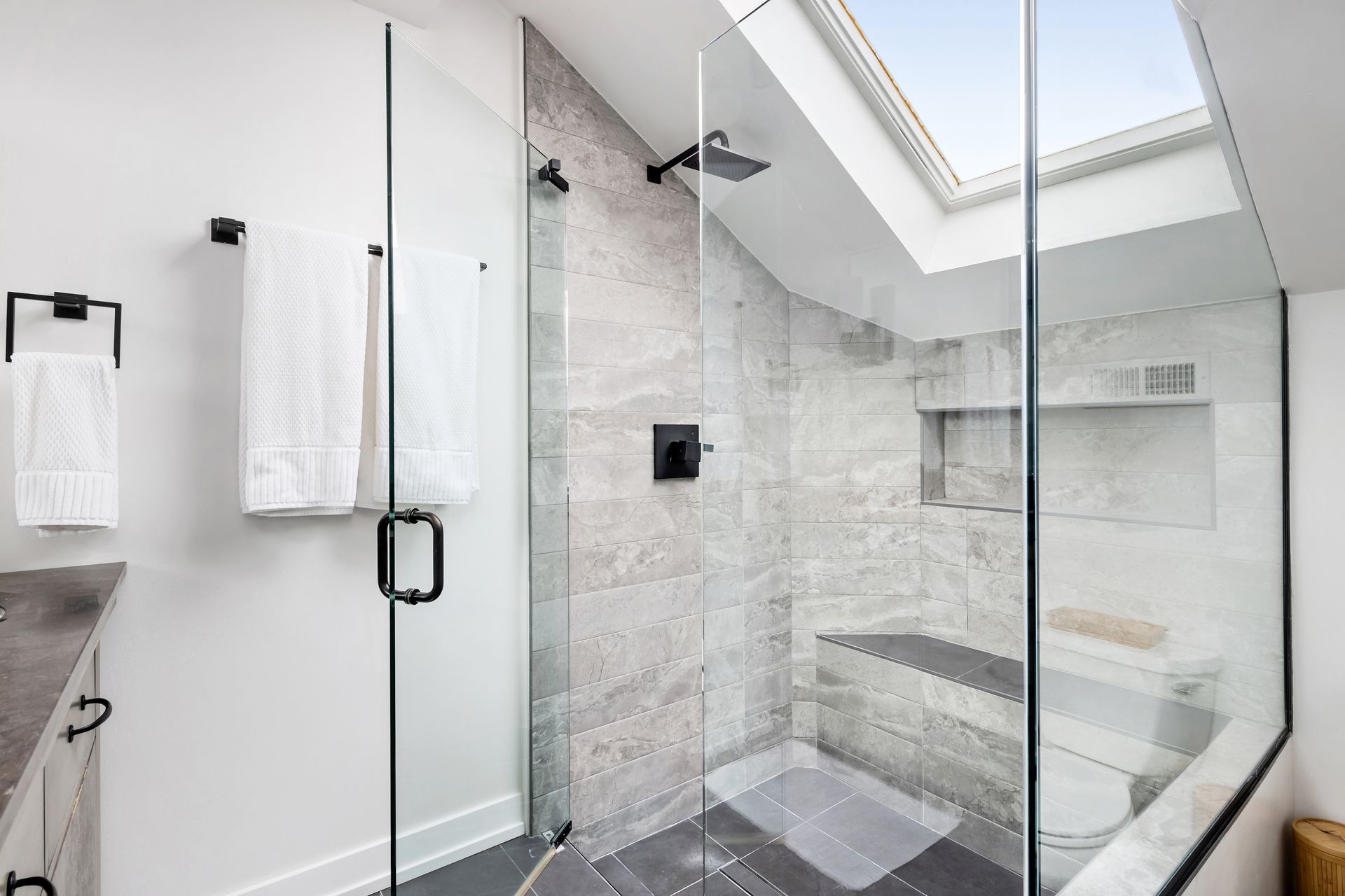 Modern walk-in shower with gray stone-look tiles, a glass enclosure, skylight, and black fixtures in a bright bathroom.