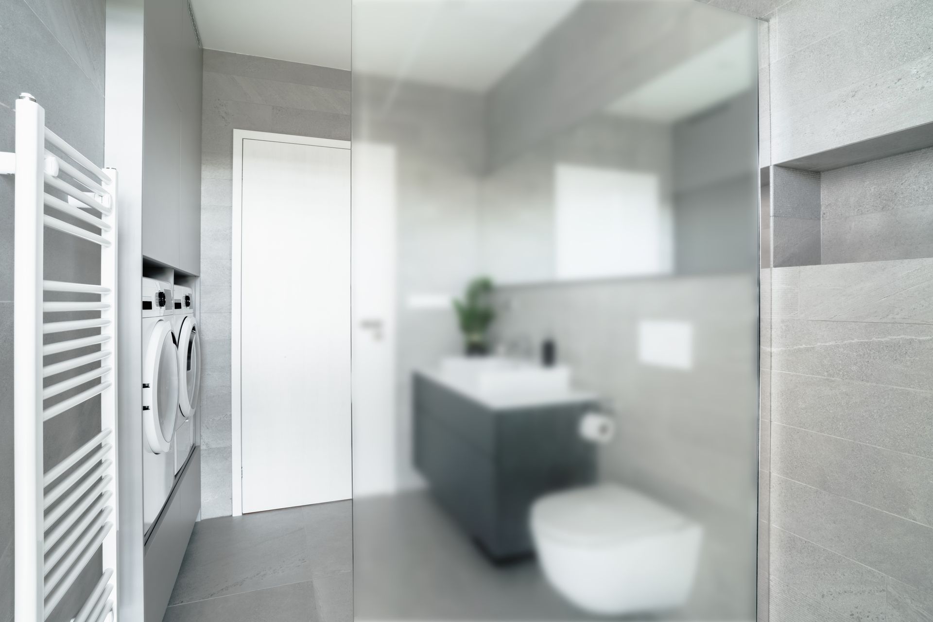 A modern, gray-tiled bathroom featuring a white heated towel rack, laundry appliances, a vanity, and a wall-hung toilet.