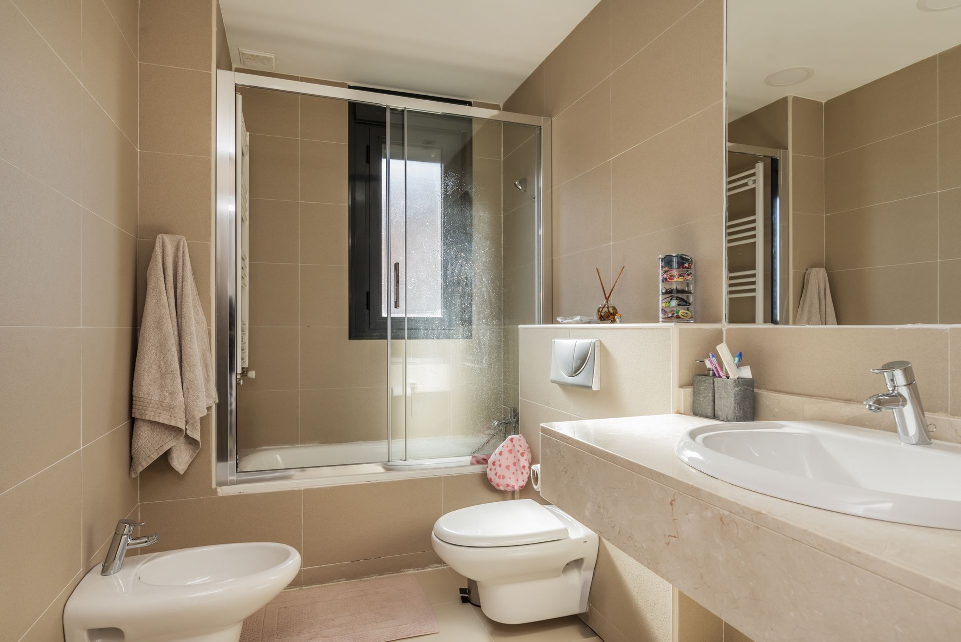 A neutral-toned bathroom with beige tiles, a bidet, a toilet, a vanity sink, and a glass-enclosed shower/tub combo.