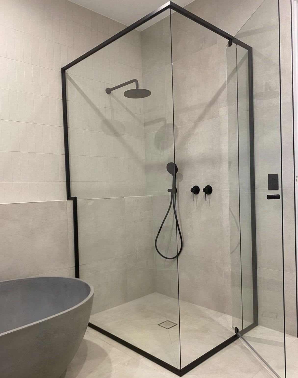 A glass-enclosed shower with matte black fixtures, white tiled walls, and a dark grey tiled floor next to a white toilet.