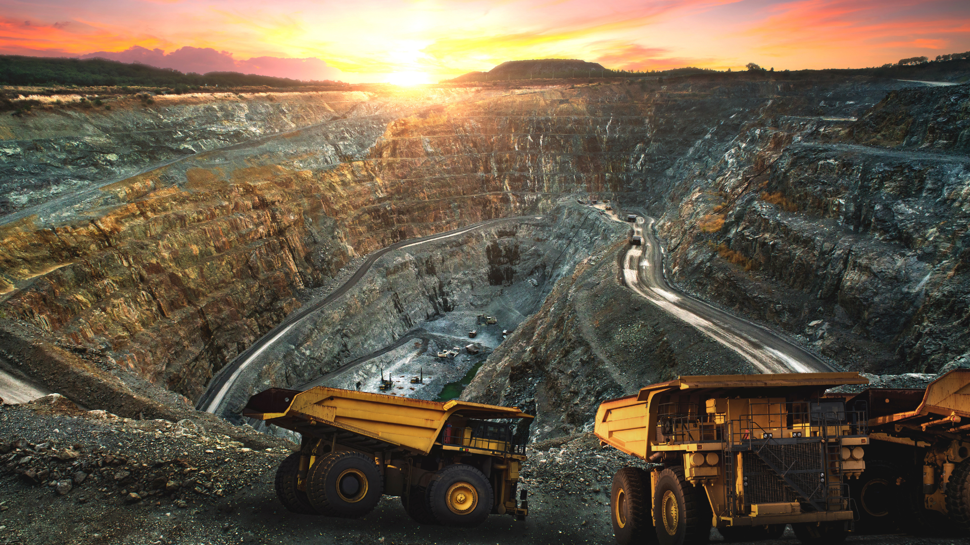 Two dump trucks are parked in front of a large open pit mine at sunset.