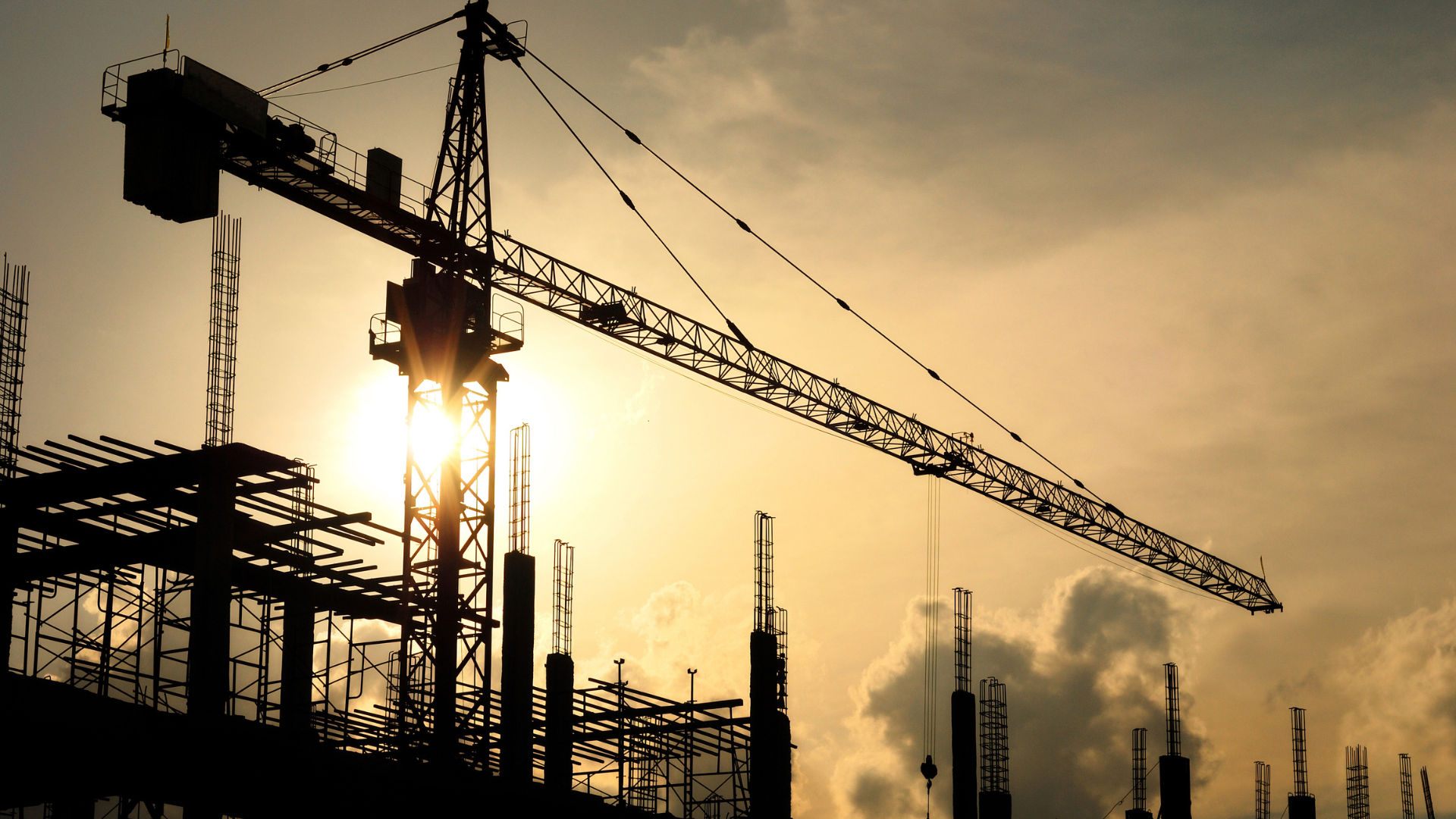A construction site with a crane in the foreground and the sun shining through the clouds