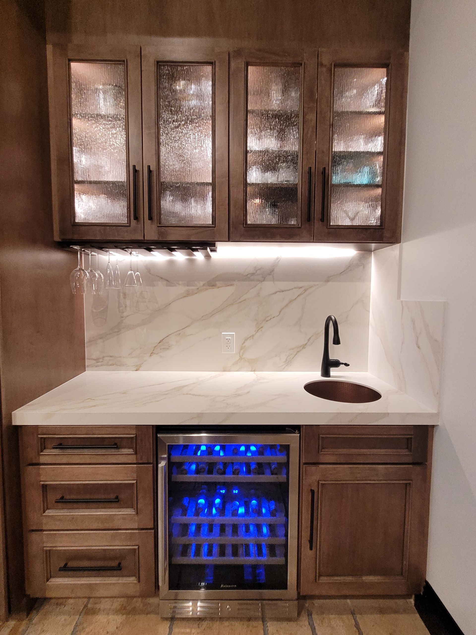 Compact wet bar with wood cabinets, white marble counter, glass-front uppers, sink, and blue-lit wine fridge.