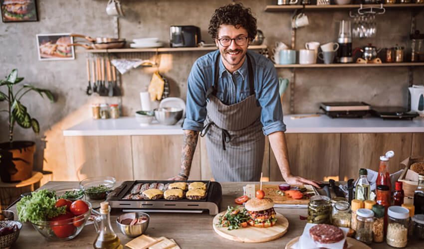 Chef smiling in a kitchen, leaning over a counter with dishes, utensils, and ingredients laid out.