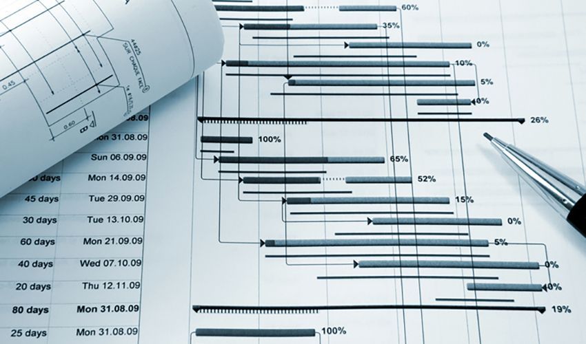 Gantt chart with task bars and percentages, over blueprints and a pen on a desk