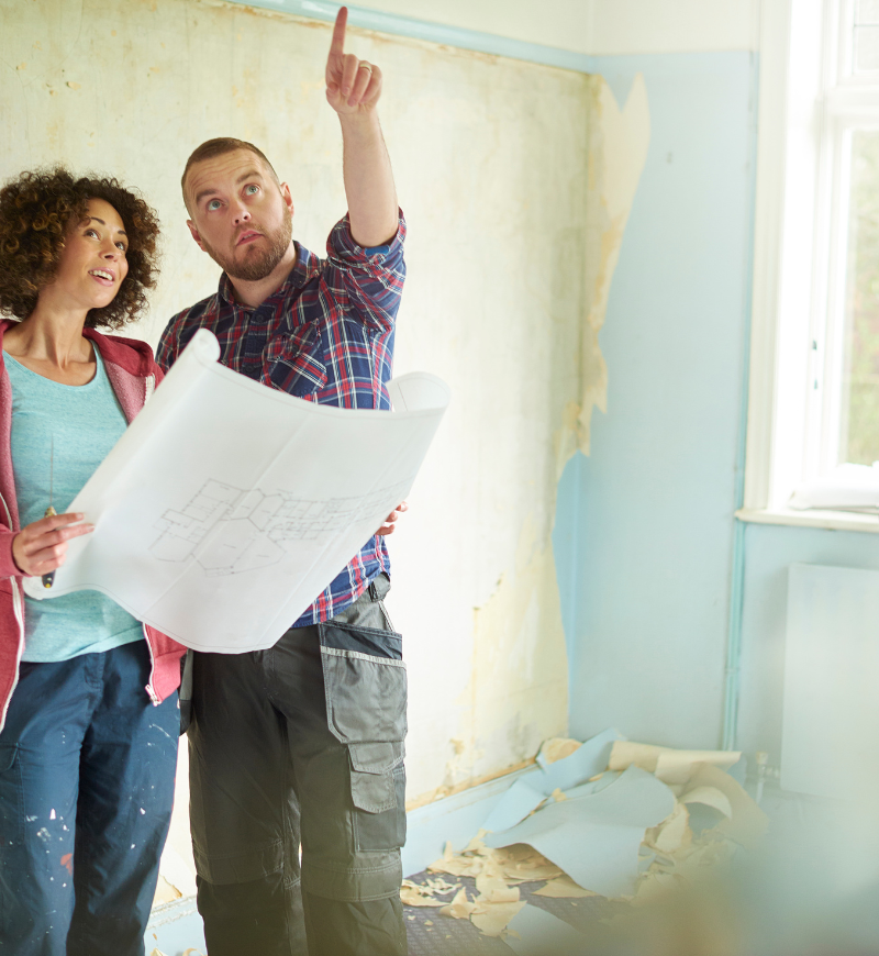 Two people reviewing a floor plan in a room under renovation, with peeling paint and construction debris.