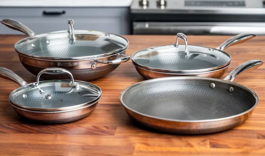 Five stainless steel frying pans and saucepans displayed on a wooden countertop in a kitchen
