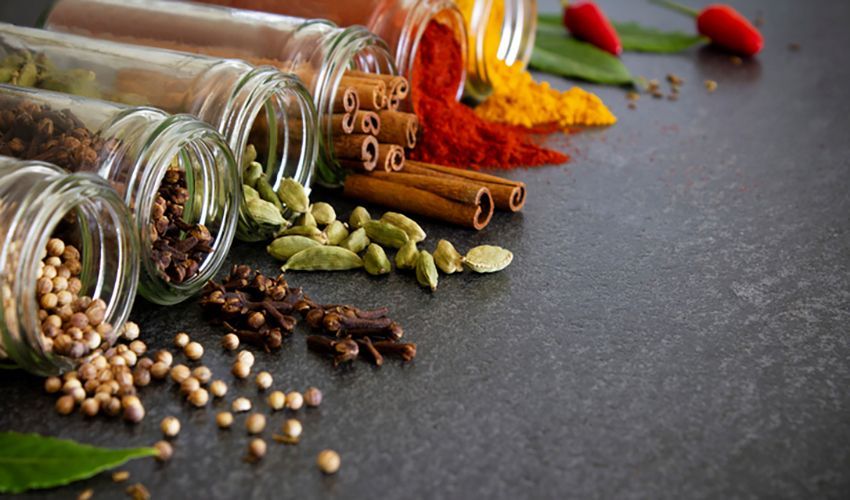 Spilled jars of colorful spices and herbs on a dark countertop, with cinnamon sticks and whole peppercorns.