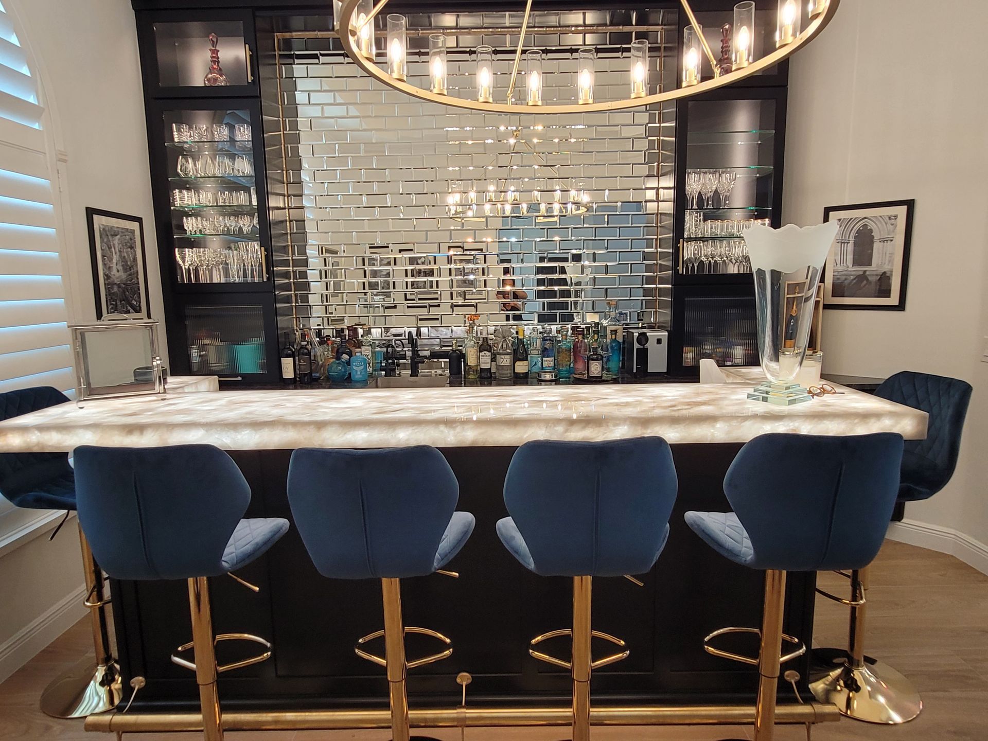 Modern bar interior with a marble counter, blue stools, mirrored back wall, and hanging light fixture