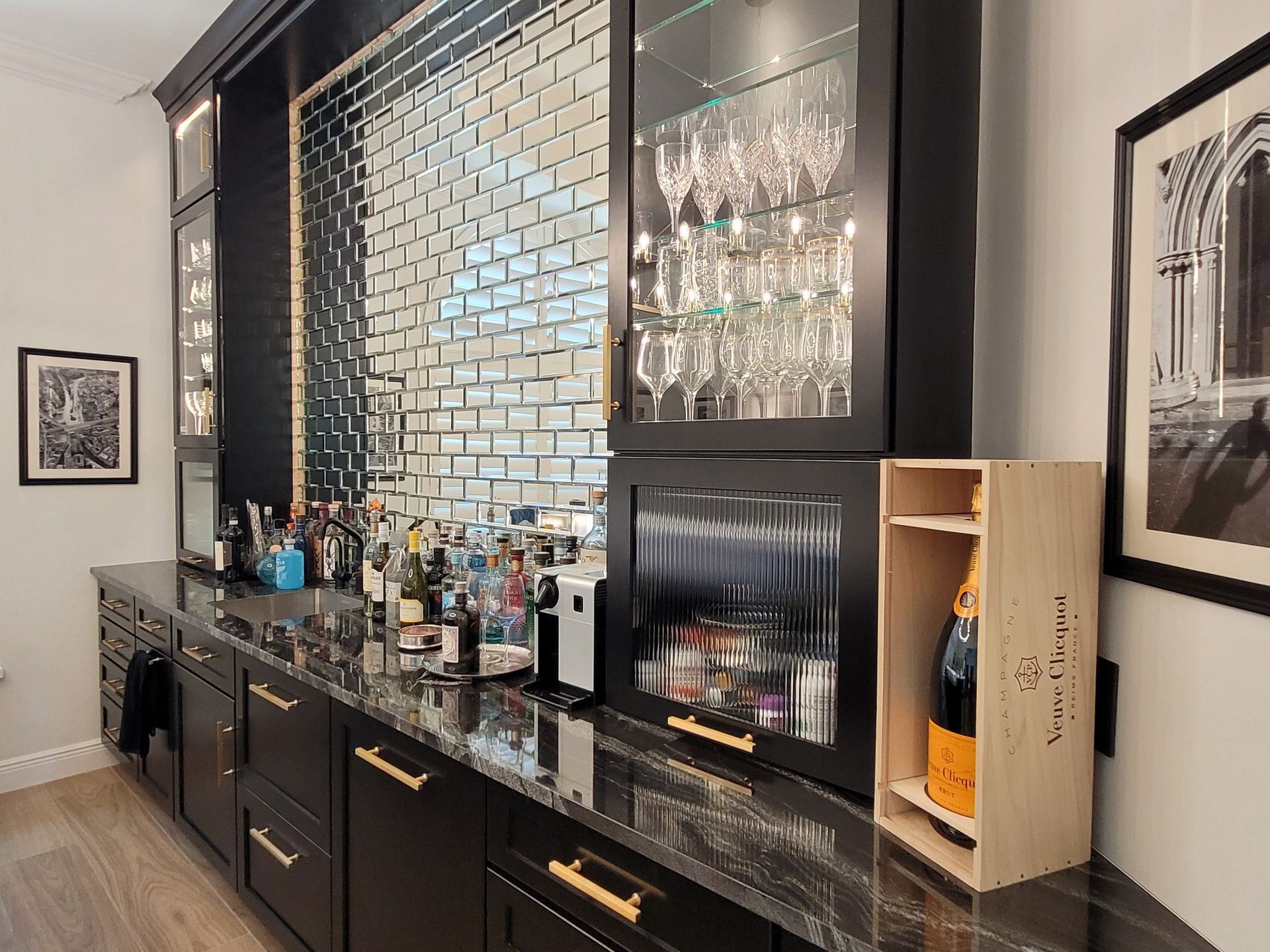 Modern home bar with mirrored tile backsplash, dark cabinetry, bottles, and framed wall art