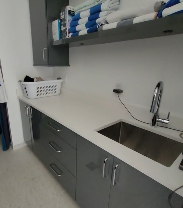 Small break room with gray cabinets, sink, countertop, and neatly stacked towels on a shelf.