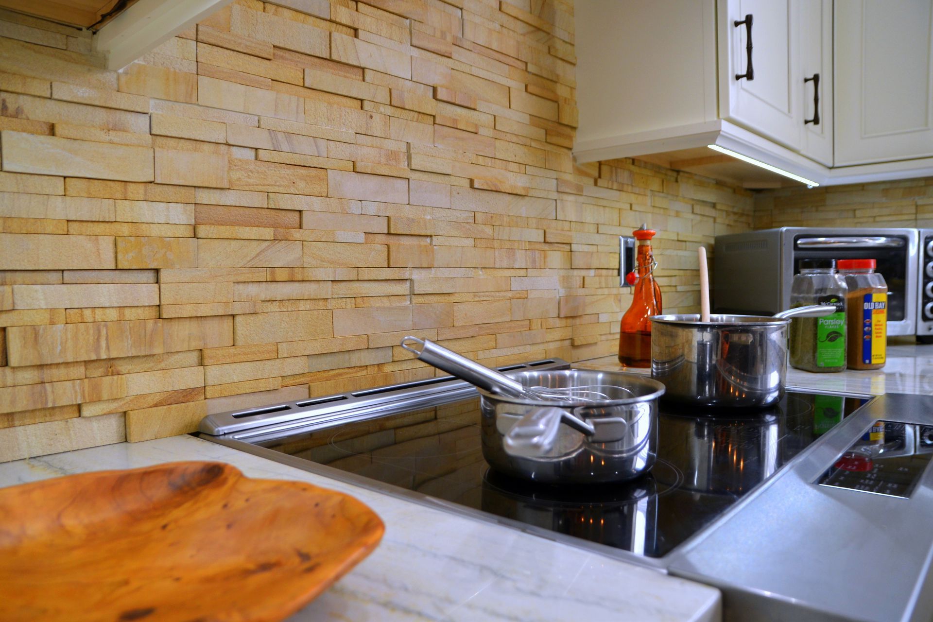 Modern kitchen counter with pots on a stove, a bottle, and a wooden cutting board with stone backsplash