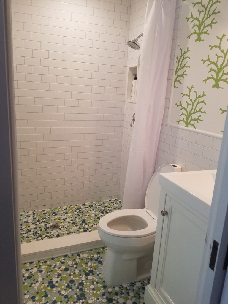 Small bathroom with white tiled shower, stone floor, toilet, sink, and green patterned wallpaper.