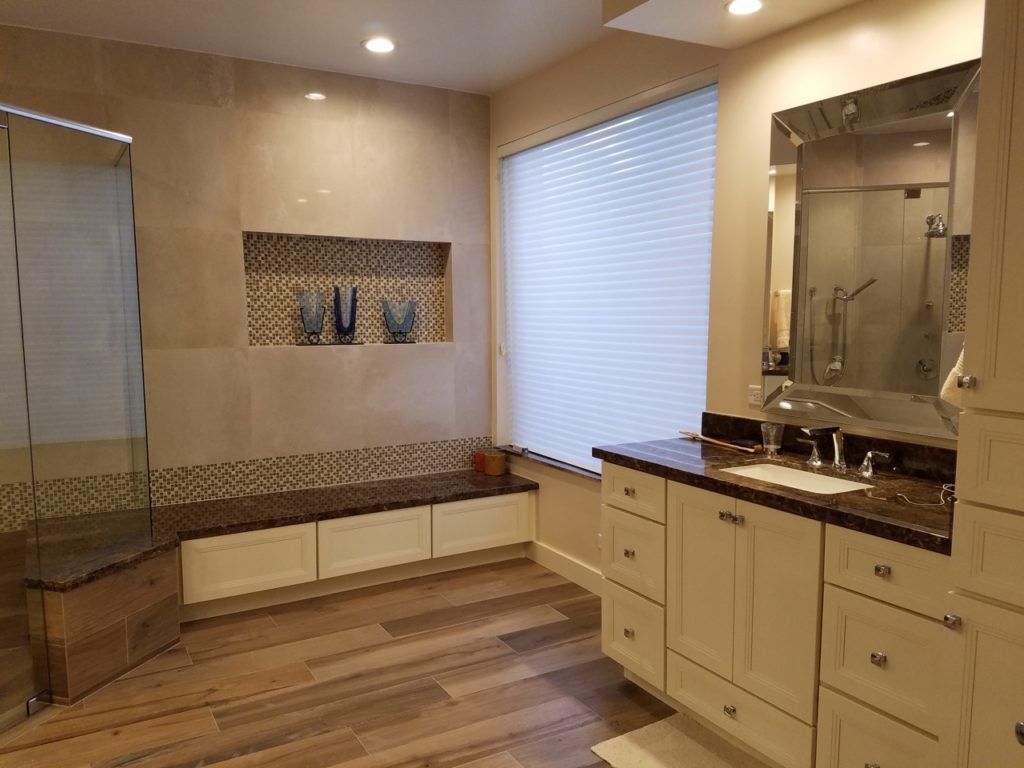 Bathroom with a wooden floor, white cabinets, brown countertop, shower, and a window with blinds.