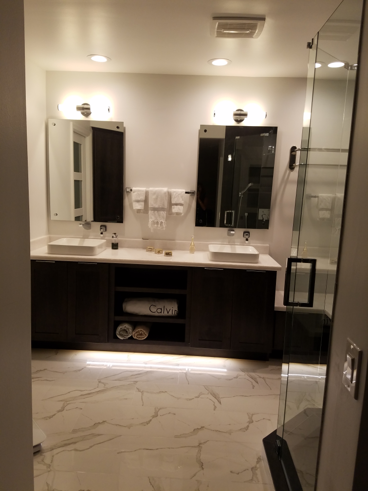 Bathroom with double sinks, dark cabinets, two mirrors, white countertop, and glass shower.