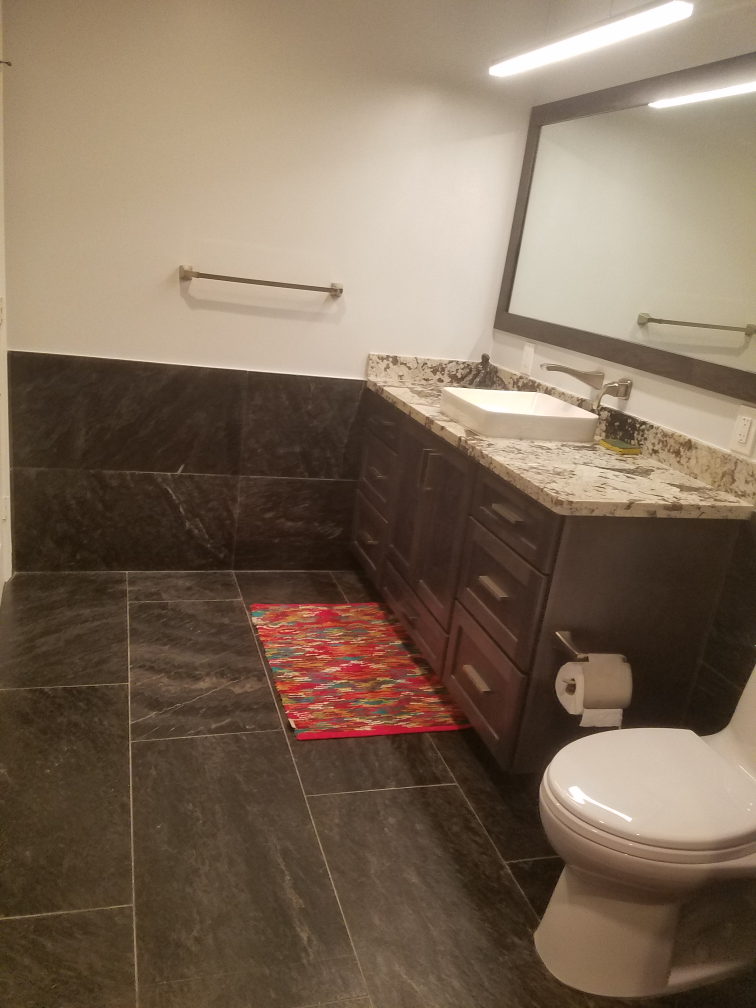 Bathroom with dark tile floor and walls, brown vanity, rectangular mirror, and red rug.