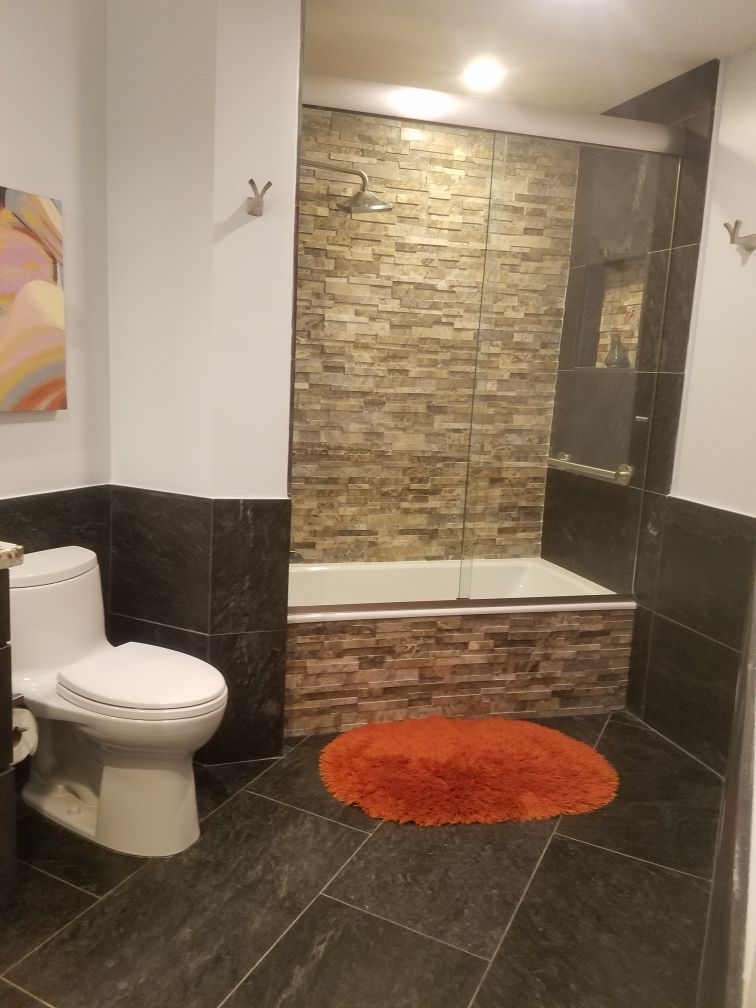Bathroom with stone tile, white toilet, orange bath mat, and glass shower door.