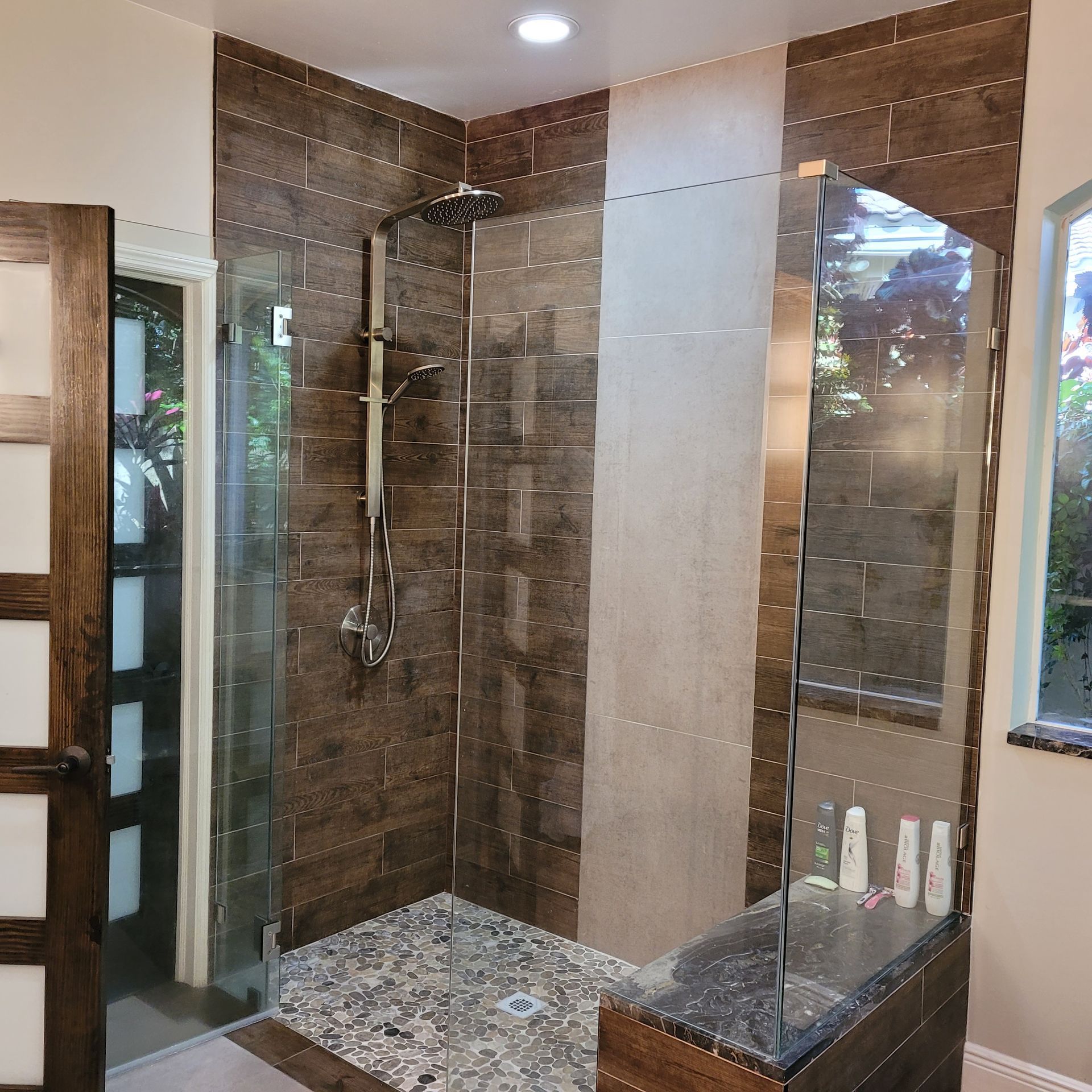 Modern shower with brown tile walls and pebble floor. Glass door and bench.