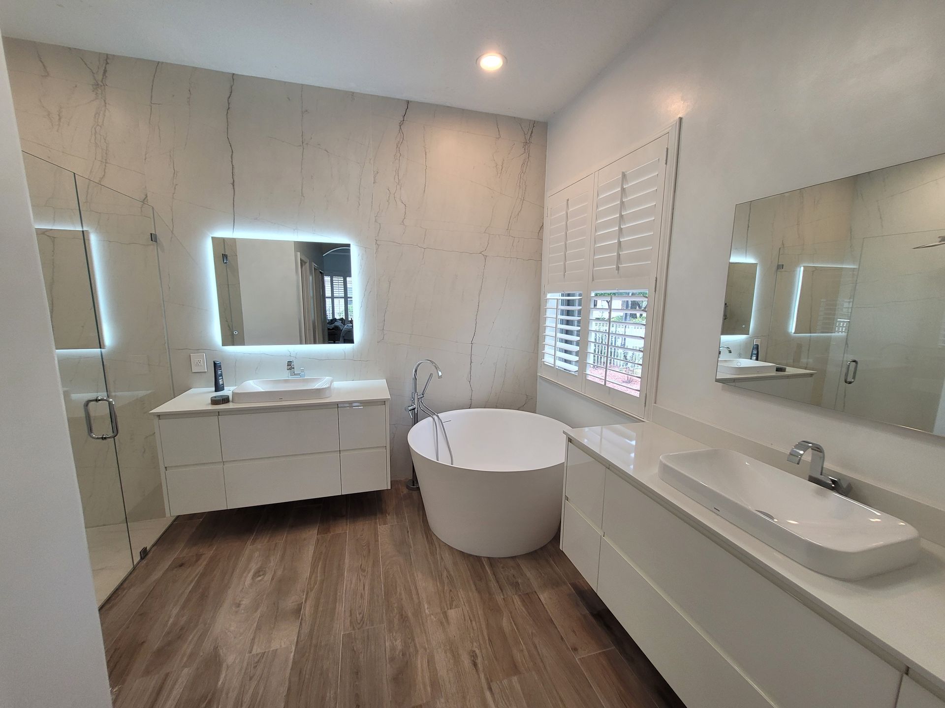 Modern bathroom with white cabinets, marble walls, wooden floor, and a freestanding tub.